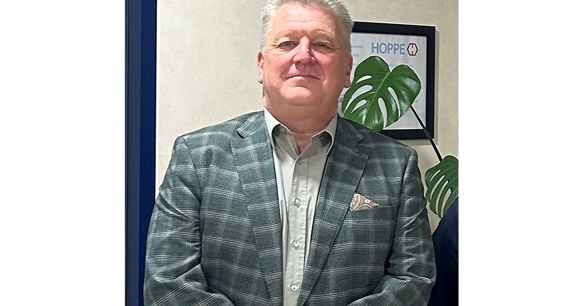 Andy Matthews standing in front of an office wall with a framed HOPPE logo, wearing a grey check blazer